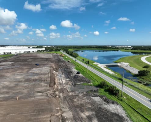Construction site for site development and land preparation with cranes, machinery, and cleared land near a water body and under a blue sky.