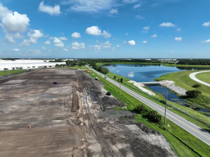Construction site for site development and land preparation with cranes, machinery, and cleared land near a water body and under a blue sky.