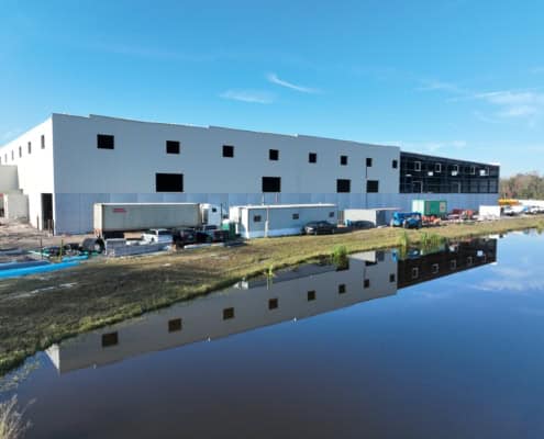Modern industrial warehouse under construction with construction site equipment and vehicles, situated beside a calm water body, representing commercial development.