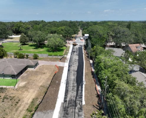 Asphalt road construction in progress with heavy machinery and safety cones, surrounded by residential homes and lush greenery, showcasing professional site development services.