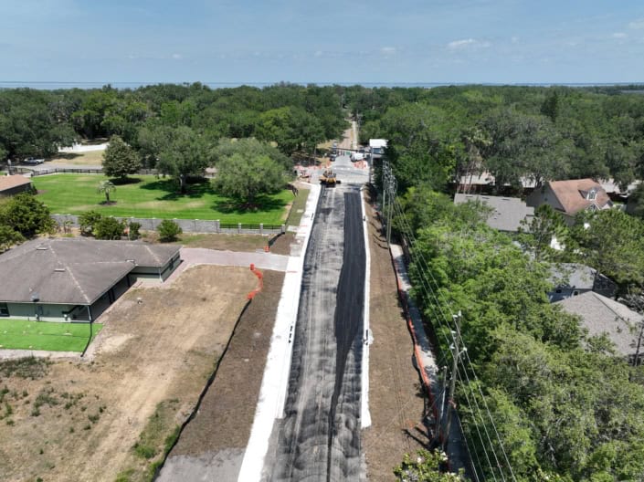 Asphalt road construction in progress with heavy machinery and safety cones, surrounded by residential homes and lush greenery, showcasing professional site development services.