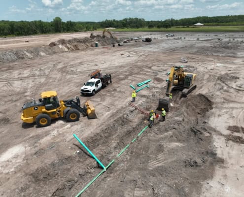 Heavy construction site with excavators, bulldozer, and workers installing utility pipes at a large development project.