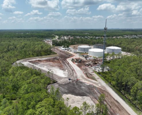Construction site of a new industrial or commercial development with storage tanks and groundwork underway, surrounded by greenery, under a partly cloudy sky.