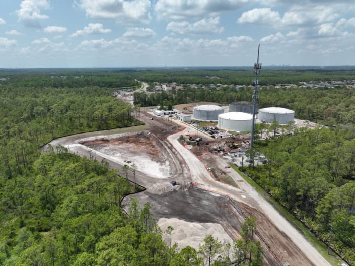 Construction site of a new industrial or commercial development with storage tanks and groundwork underway, surrounded by greenery, under a partly cloudy sky.