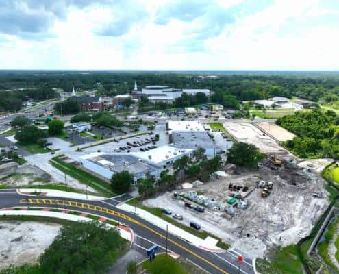 Aerial view of commercial site development with ongoing construction, including parking lots, roads, and green spaces, in a thriving community.