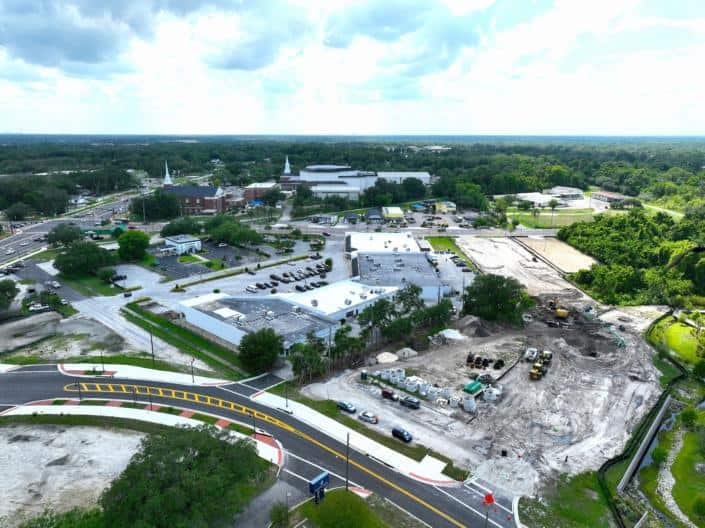 Aerial view of commercial site development with ongoing construction, including parking lots, roads, and green spaces, in a thriving community.