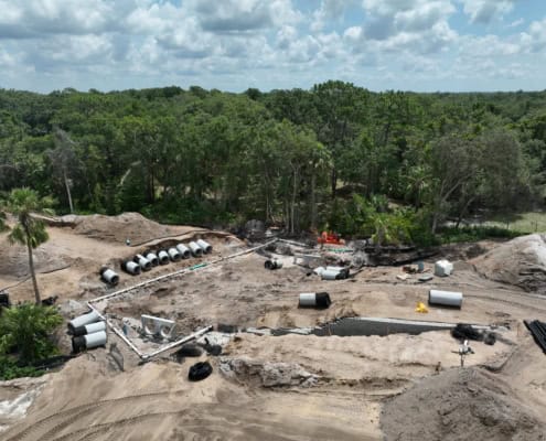 Underground construction site for site development with pipes, heavy machinery, and cleared land, surrounded by dense green trees, under partly cloudy sky.