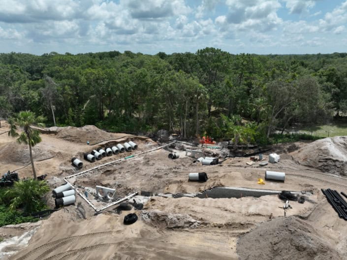 Underground construction site for site development with pipes, heavy machinery, and cleared land, surrounded by dense green trees, under partly cloudy sky.