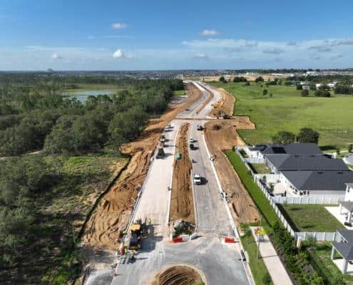New residential street under construction with paving, machinery, and workers, surrounded by green landscapes, trees, and modern houses, showing site development progress.