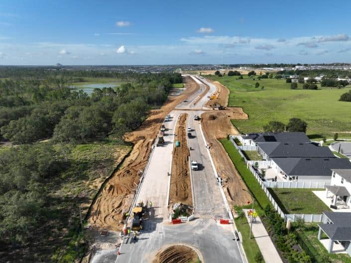 New residential street under construction with paving, machinery, and workers, surrounded by green landscapes, trees, and modern houses, showing site development progress.