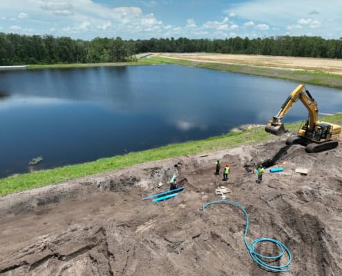Excavation work at a construction site near a large water body with an excavator and workers in safety vests, focusing on site development and land preparation for development projects.