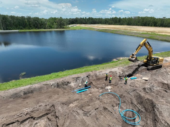 Excavation work at a construction site near a large water body with an excavator and workers in safety vests, focusing on site development and land preparation for development projects.