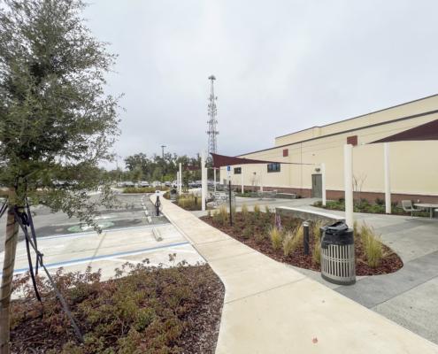 Modern commercial building exterior with landscaped surroundings, outdoor seating, and electric vehicle charging station.