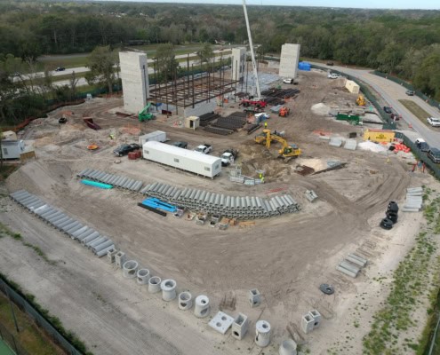 Heavy construction site with building framework, large pipes, construction machinery, and workers at a commercial development project.