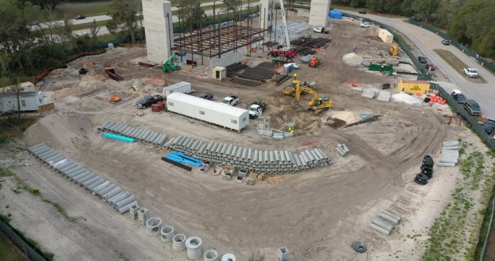 Heavy construction site with building framework, large pipes, construction machinery, and workers at a commercial development project.
