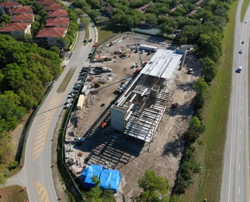 Modern commercial building under construction with steel framework and ongoing roofing work, located in a rapidly developing urban area with nearby residential neighborhoods and roads.