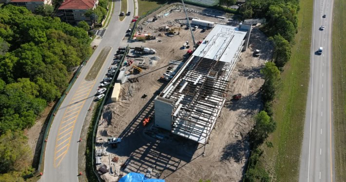 Modern commercial building under construction with steel framework and ongoing roofing work, located in a rapidly developing urban area with nearby residential neighborhoods and roads.