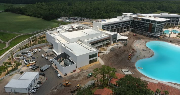 Modern hotel construction site with a large swimming pool, surrounded by greenery and ongoing building activities, showcasing contemporary architecture and real estate development.